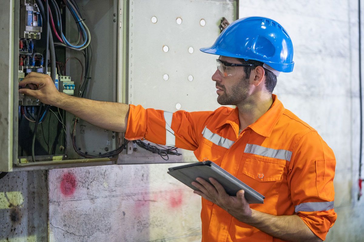 Electrician in orange uniform and blue hard hat inspects a control panel, holding a tablet.