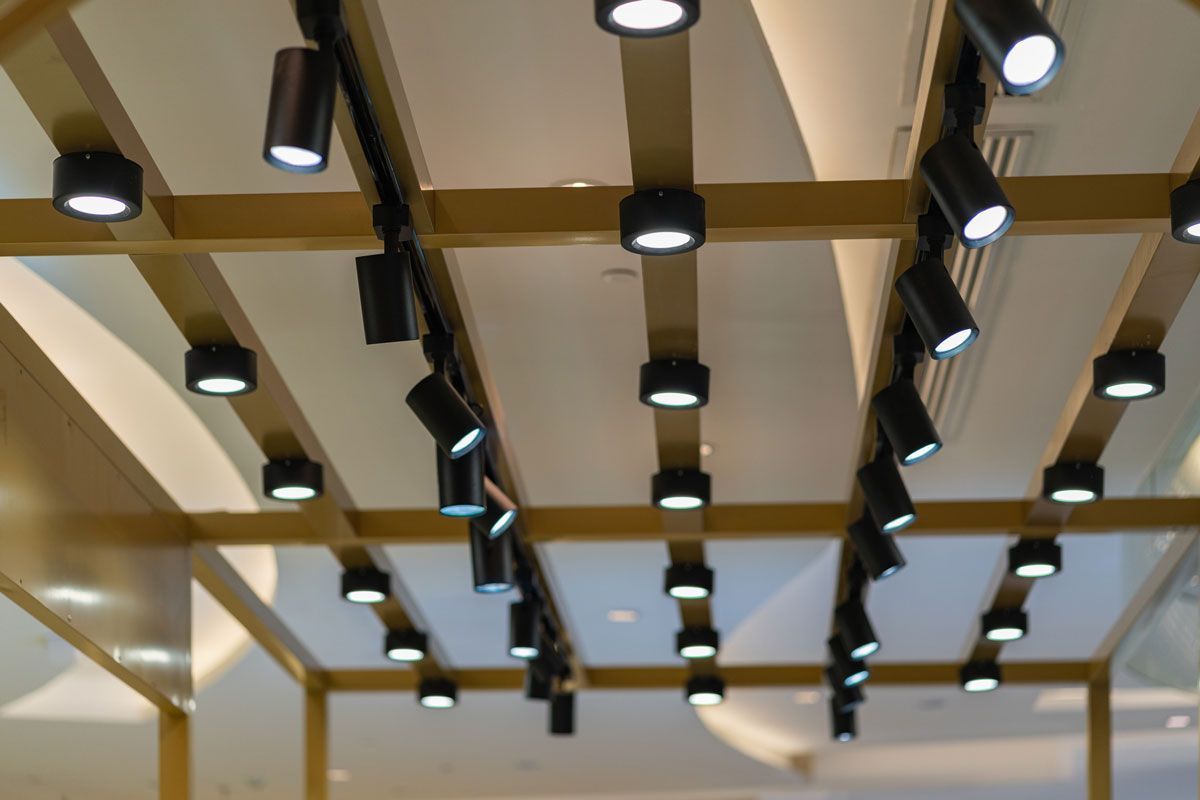 Black track lighting fixtures mounted on a gold grid ceiling, illuminating a white ceiling.
