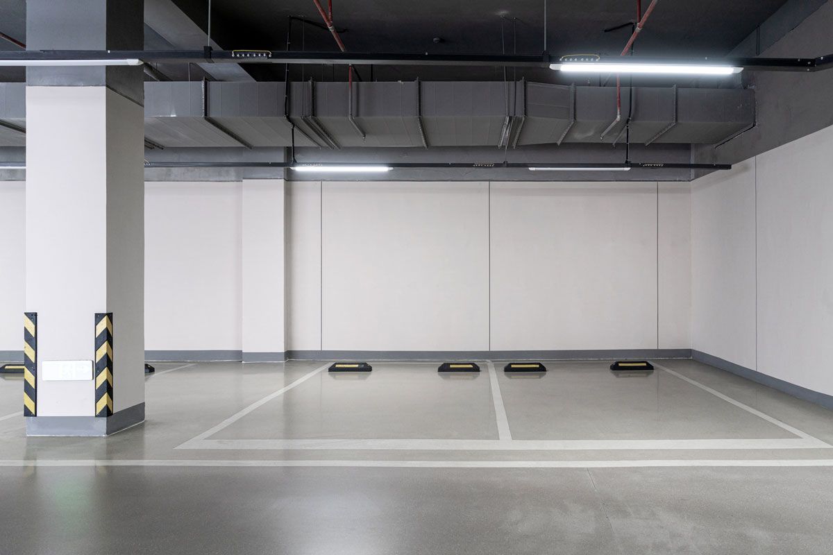 Empty parking garage with white walls, gray floor, and overhead lighting. Concrete pillars and parking space markers are visible.