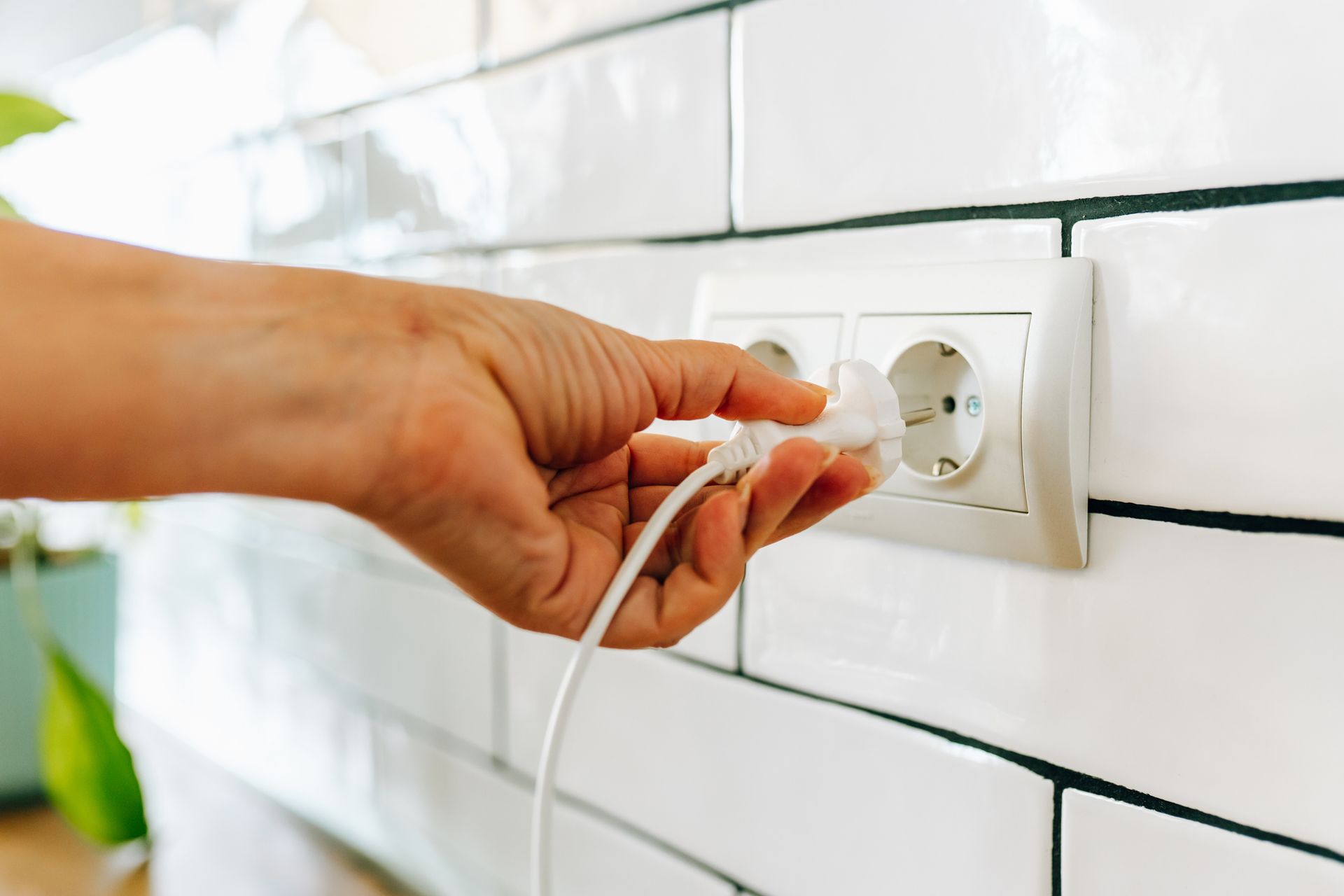 Hand plugging a white cord into a European-style wall outlet. White tiled wall in the background.