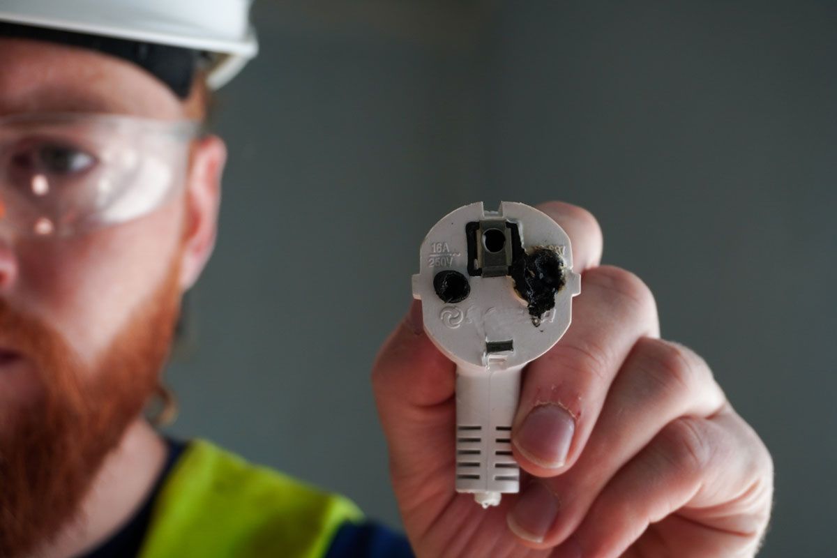 Electrician holding a damaged white electrical plug with burn marks; safety glasses and hard hat visible.