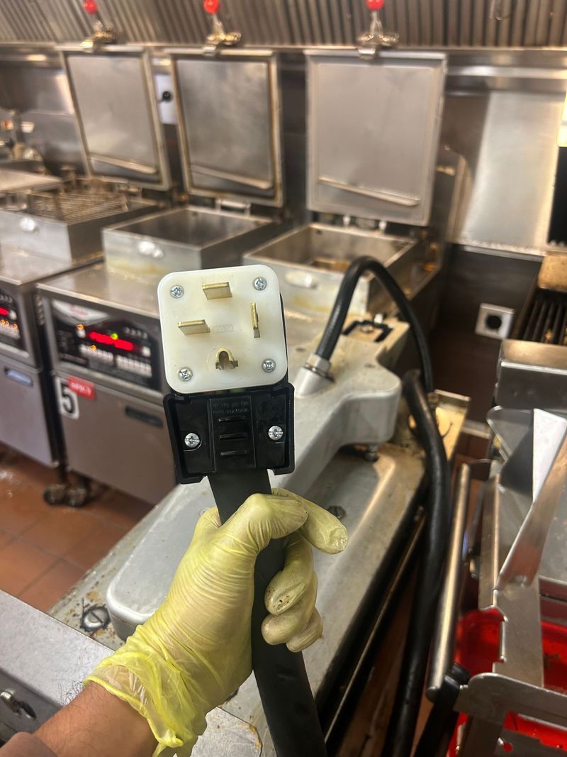 Electrician holding a damaged white electrical plug with burn marks; safety glasses and hard hat visible.