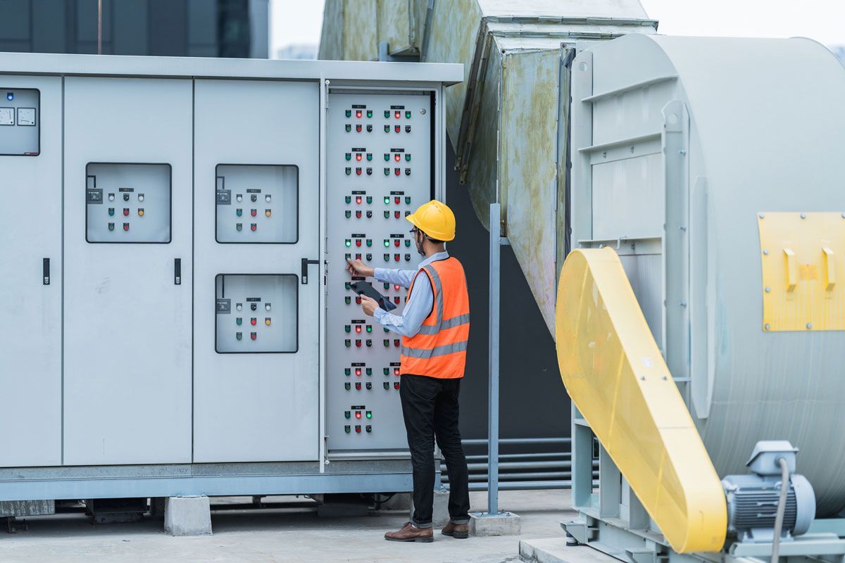 Person in safety vest and hard hat inspecting electrical panel on rooftop near industrial equipment.