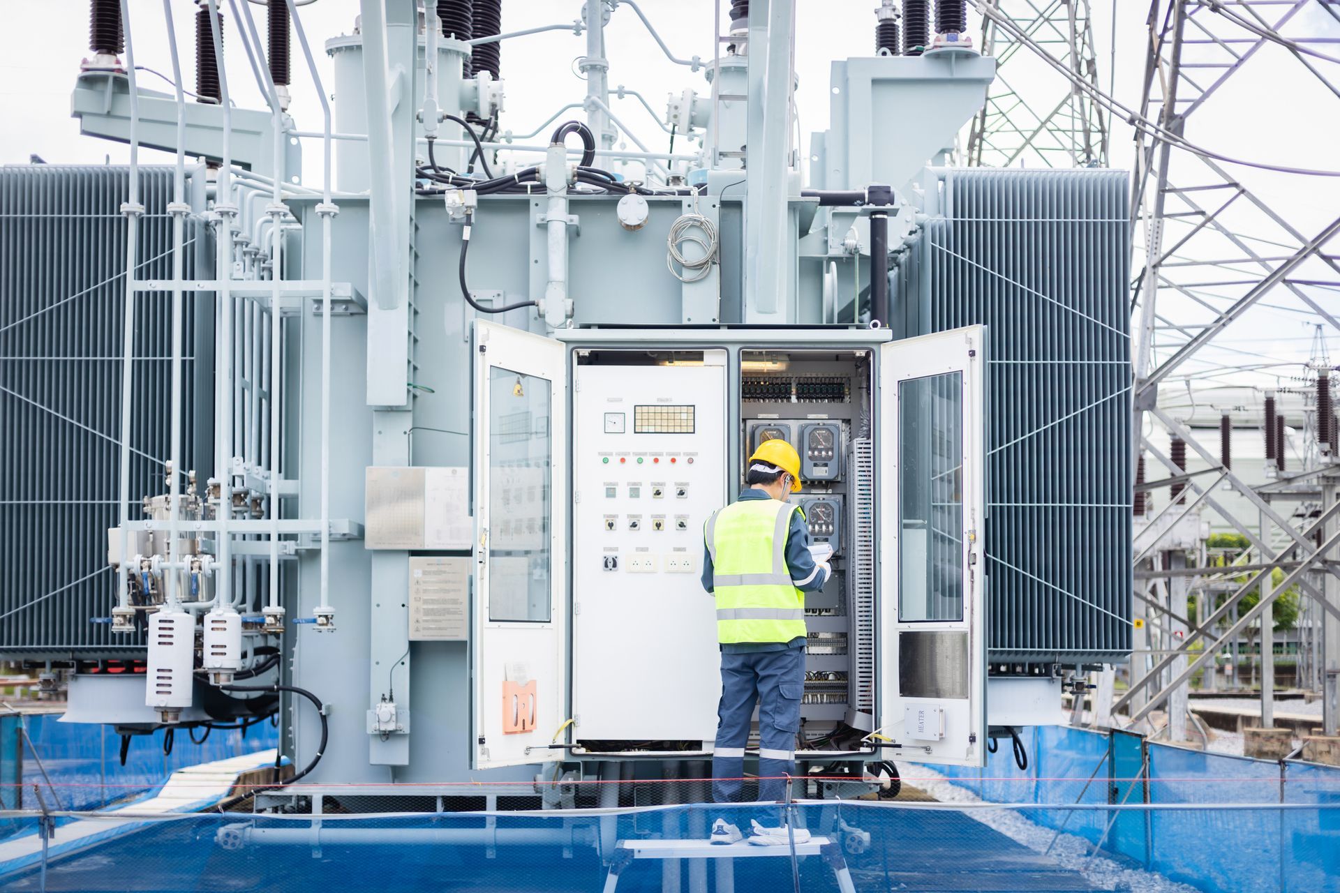 Electrician in yellow vest, inspecting control panel of a large transformer at a power station.