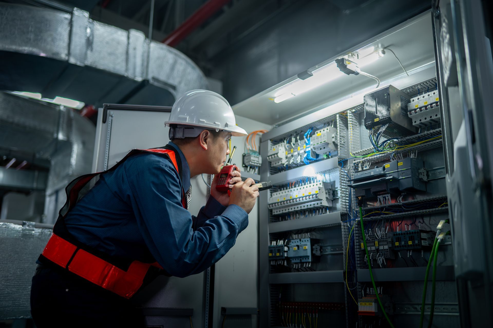 Electrician in hard hat inspecting electrical panel with a flashlight in a mechanical room.