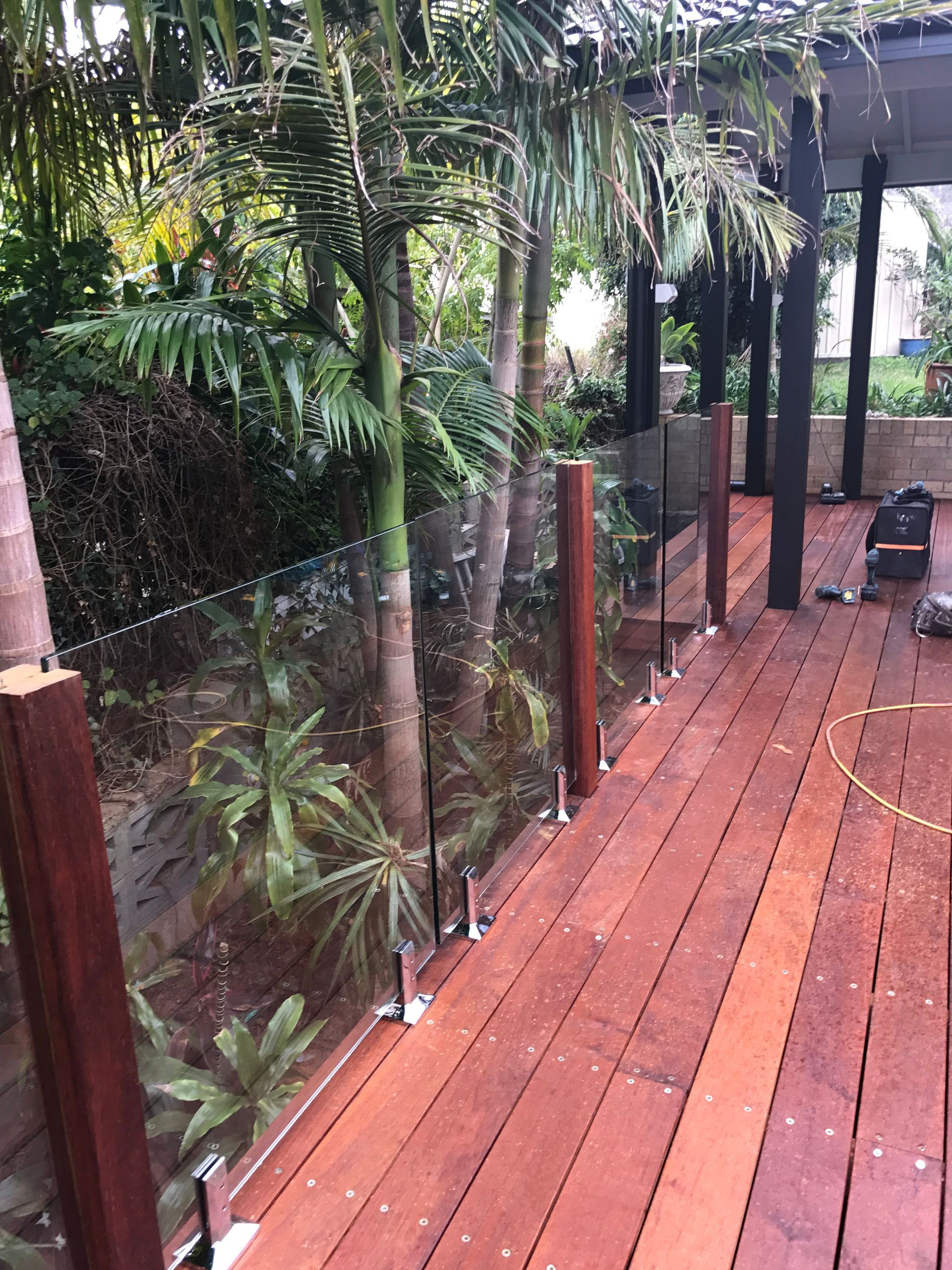 Wooden deck with glass railing bordering lush greenery and palm trees.
