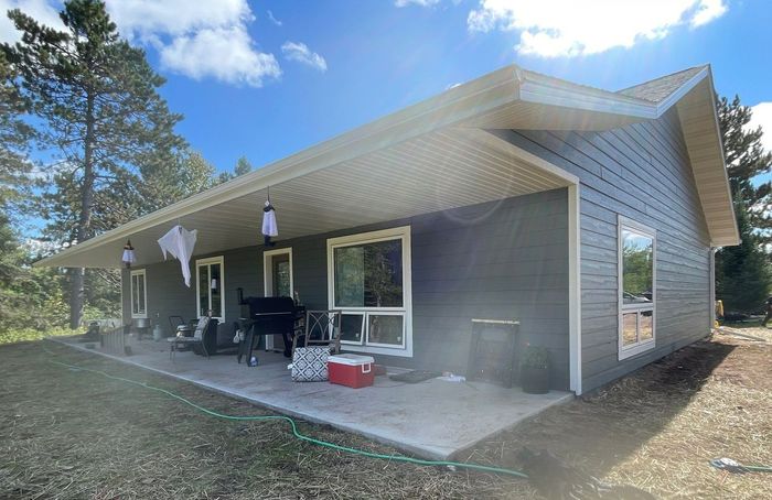 A grey, horizontal-sided house with a wide covered concrete patio under a clear blue sky with scattered clouds.