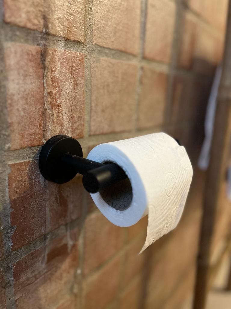 Een rol toiletpapier hangt aan een zwarte toiletrolhouder aan een bakstenen muur.