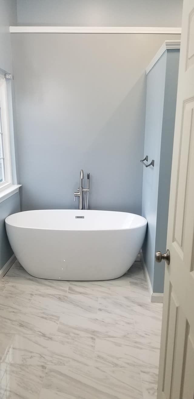 A modern white freestanding bathtub set against a pale blue wall with light-colored, wood-look tiled flooring.