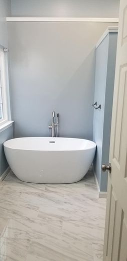 A modern white freestanding bathtub set against a pale blue wall with marble-style tile flooring in a bathroom.