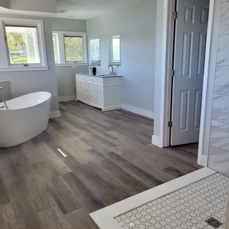 A bright modern bathroom with grey wood-look floors, a white soaking tub, a double vanity, and a tiled shower entry.