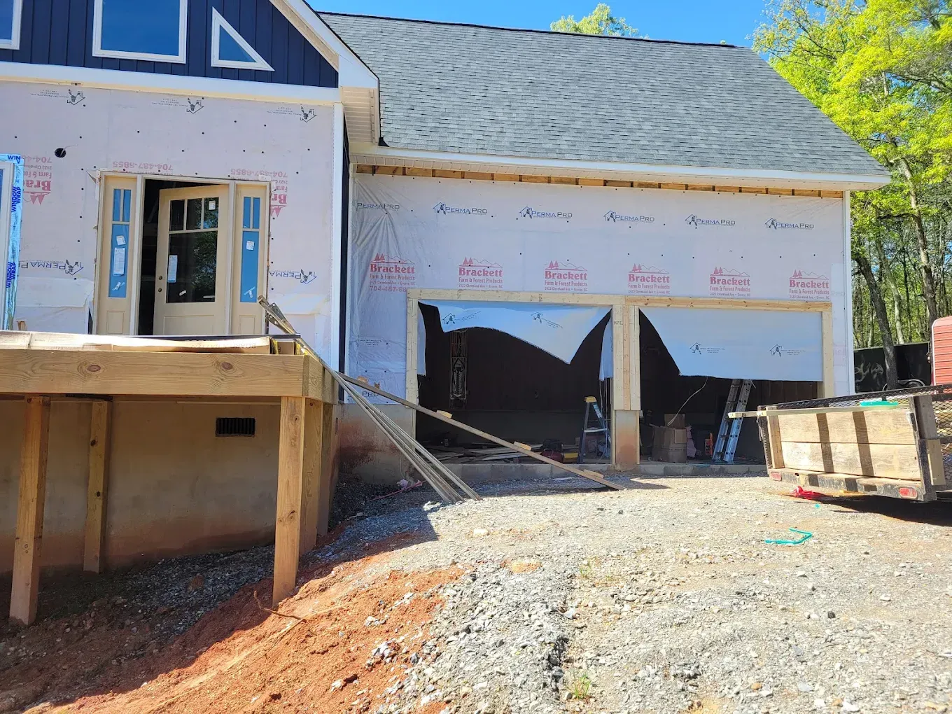 A house under construction with a wooden deck, uninstalled garage doors, and white protective house wrap on exterior walls.