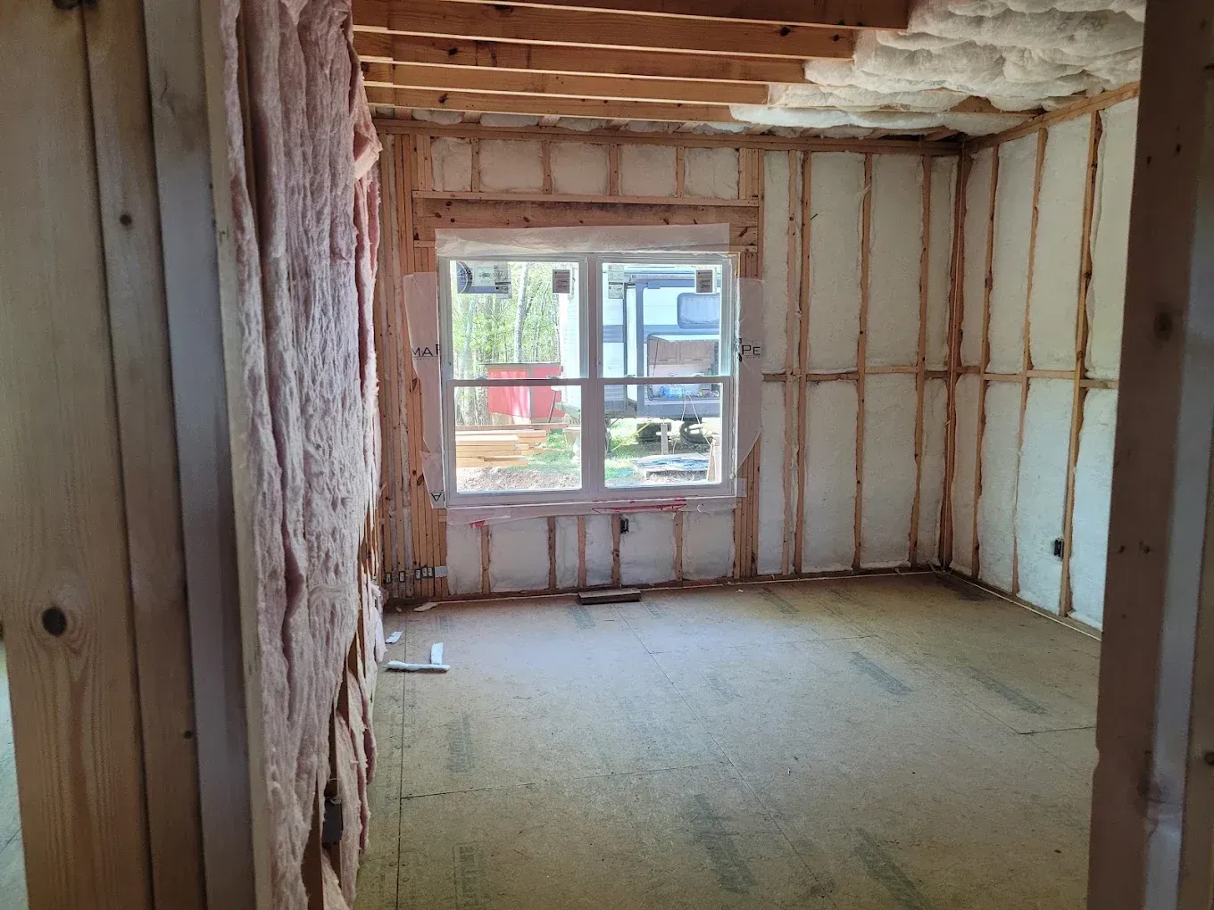 An interior view of a room under construction with exposed wooden wall studs, foam insulation, and a central window.