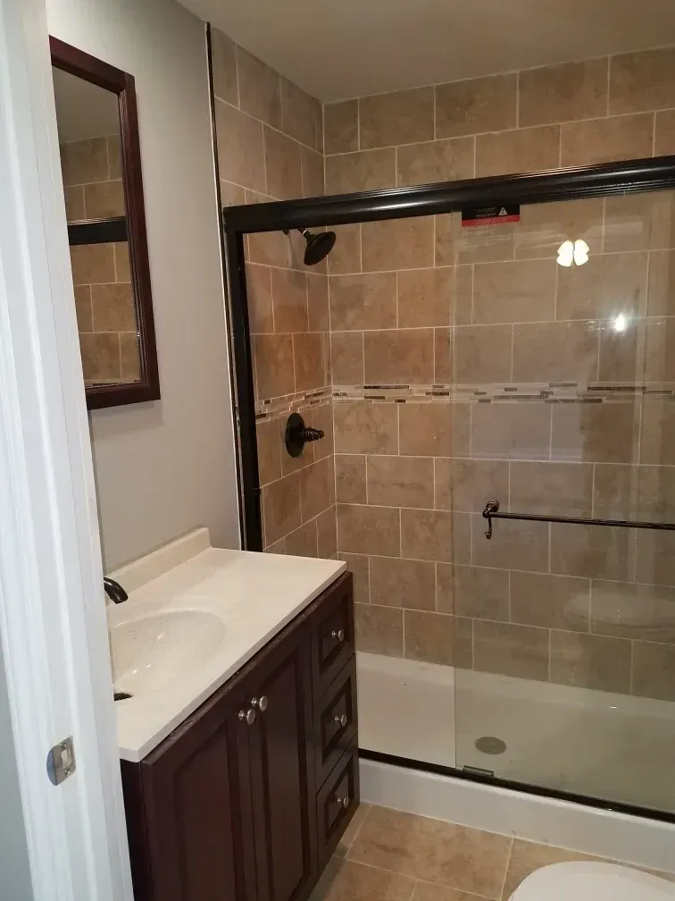 A bathroom featuring a dark wood vanity with a white countertop, a mirror, and a glass-enclosed shower with beige tiling.