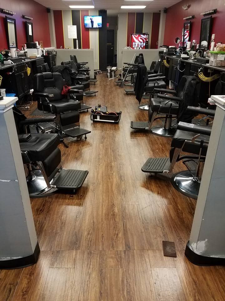 A barbershop interior with rows of black chairs facing mirrors along red walls, featuring wood-look floors.