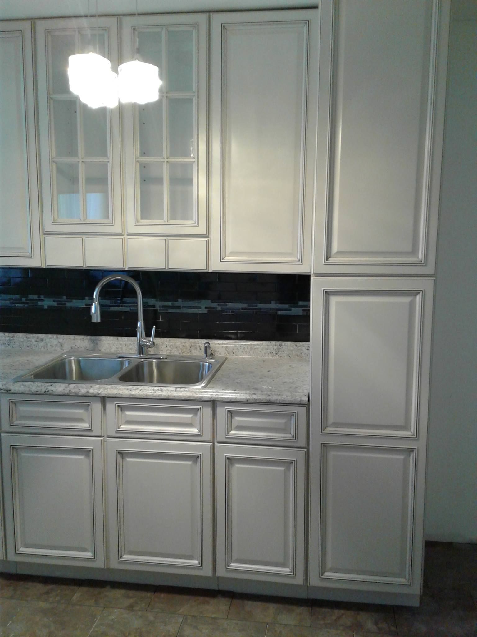 A kitchen sink area featuring grey cabinets with dark glazing, a granite countertop, and a black tiled backsplash.