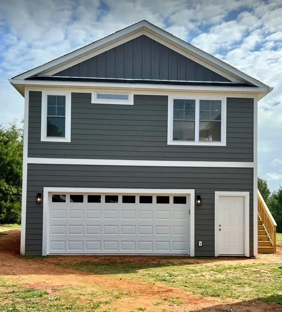 A detached two-story garage with dark gray horizontal siding, a white garage door, a side door, and exterior lights.