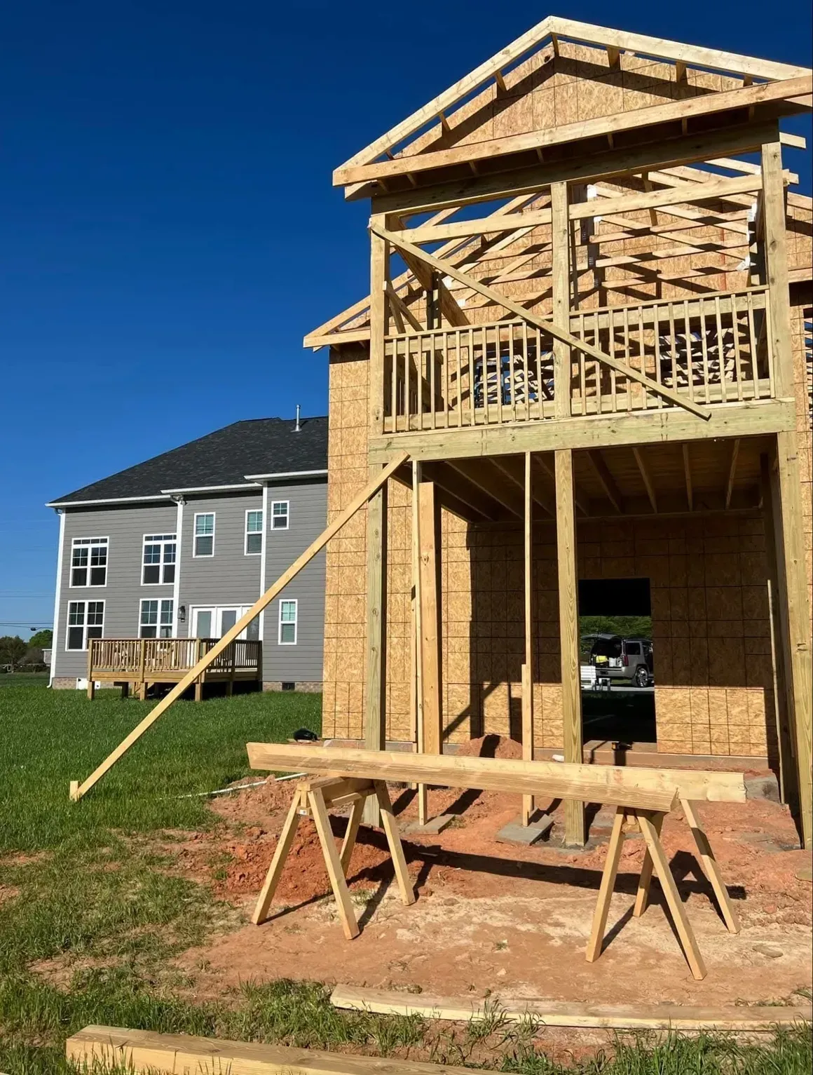 A house under construction with exposed wooden framing and a large support beam on sawhorses, next to a finished home.