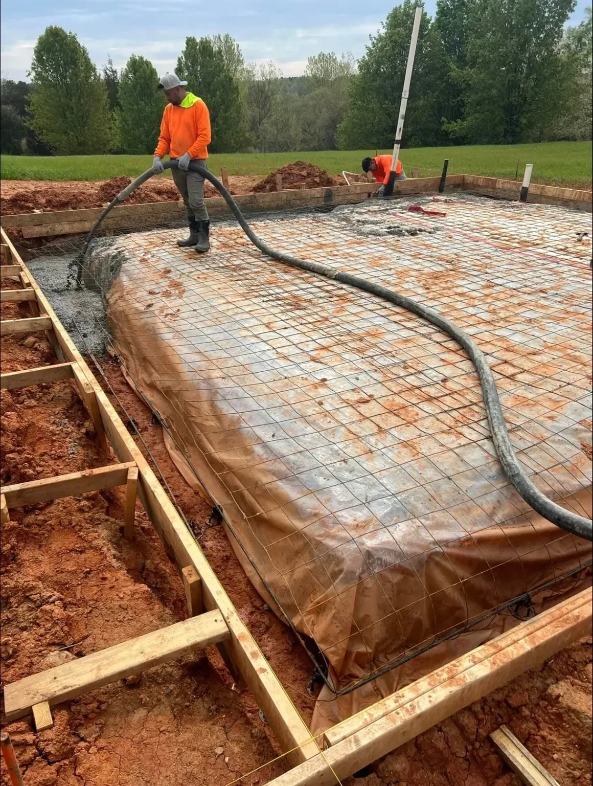 Workers in high-visibility orange jackets pouring concrete into a foundation form with a hose at a construction site.