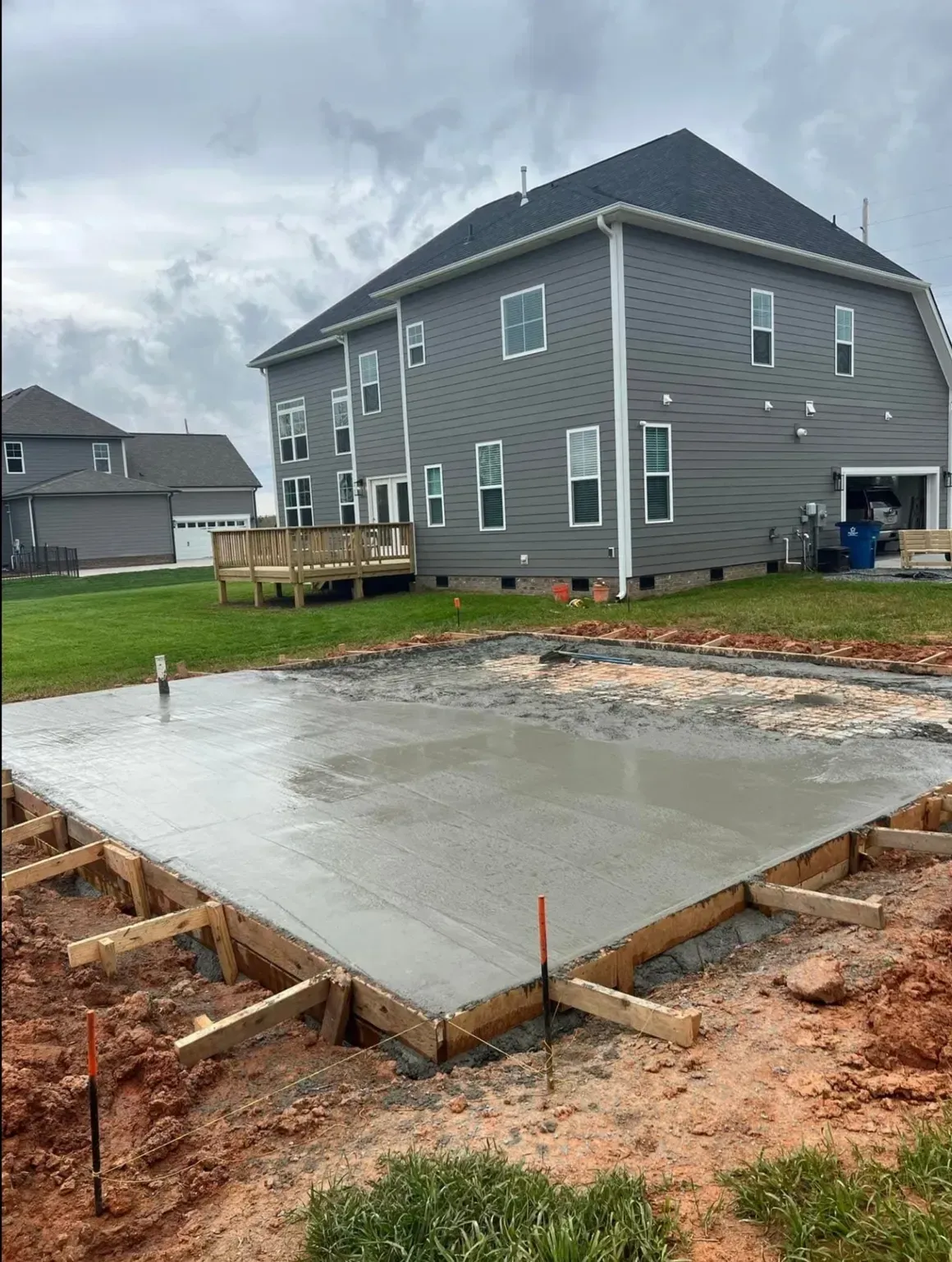 A freshly poured concrete patio slab sits in a dirt backyard next to a gray two-story house.