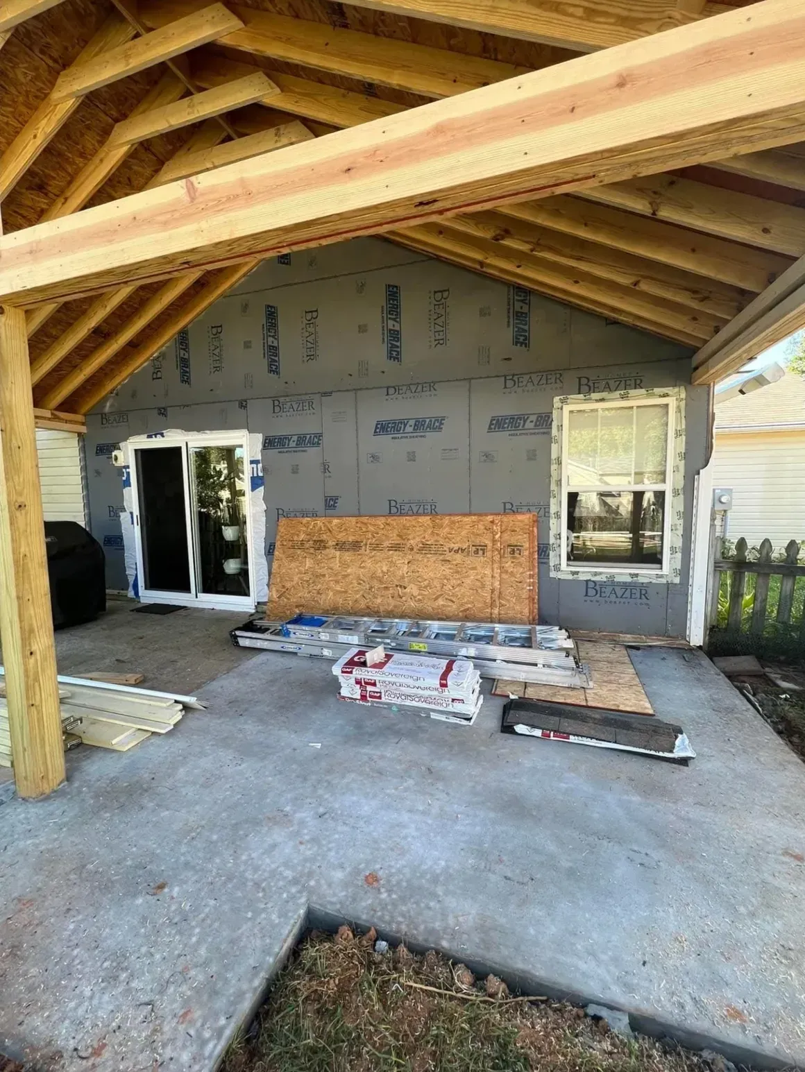 A covered patio under construction featuring wood framing, gray exterior sheathing, a glass sliding door, and a window.