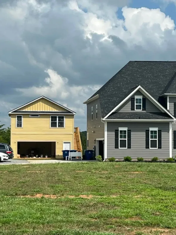 A two-story yellow garage stands beside a larger gray suburban house under a cloudy sky.