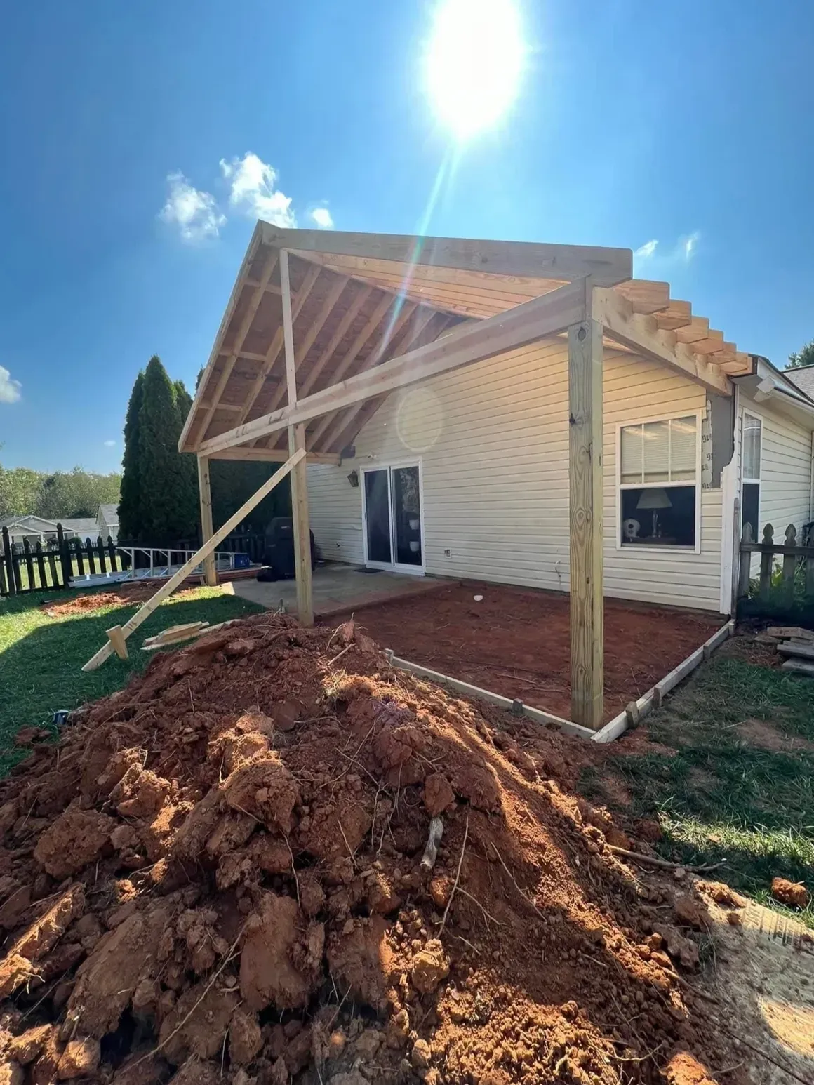 Wooden frame of a house patio addition under construction, with a large pile of dirt in the foreground.