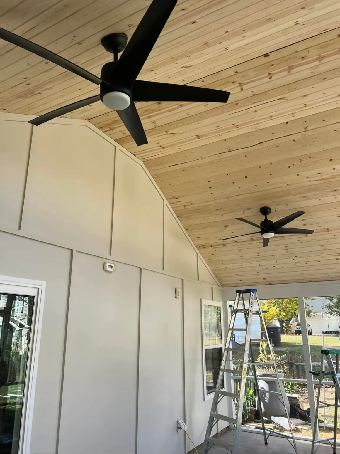 Two matte black ceiling fans with lights installed on a sloped, natural wood plank ceiling of a covered patio.