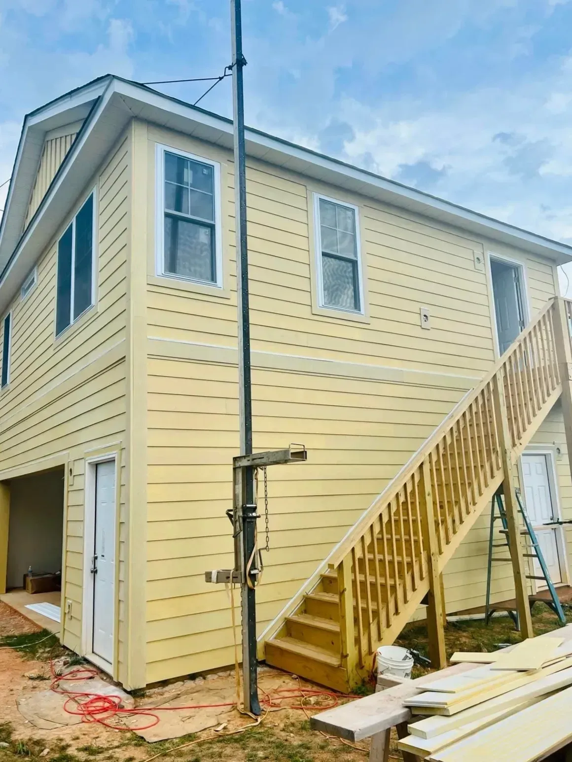 A two-story, yellow-sided house under construction with an exterior wooden staircase and a tall metal pole in the yard.