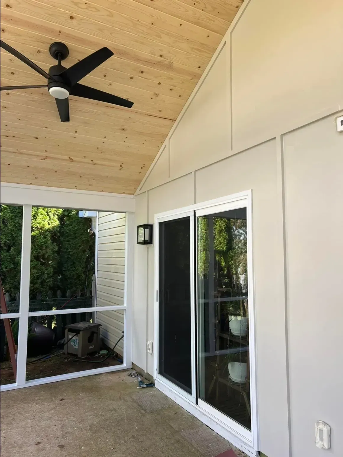 A screened-in porch with light wood ceiling, black ceiling fan, beige walls, and white sliding glass door.