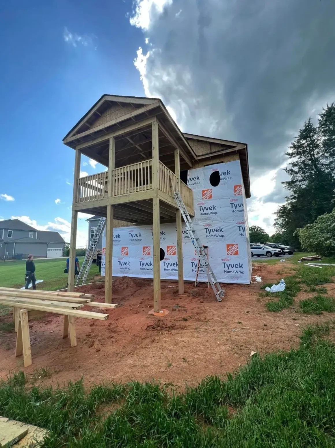 A wooden elevated deck under construction on the side of a house with Tyvek wrap on a bright, partly cloudy day.