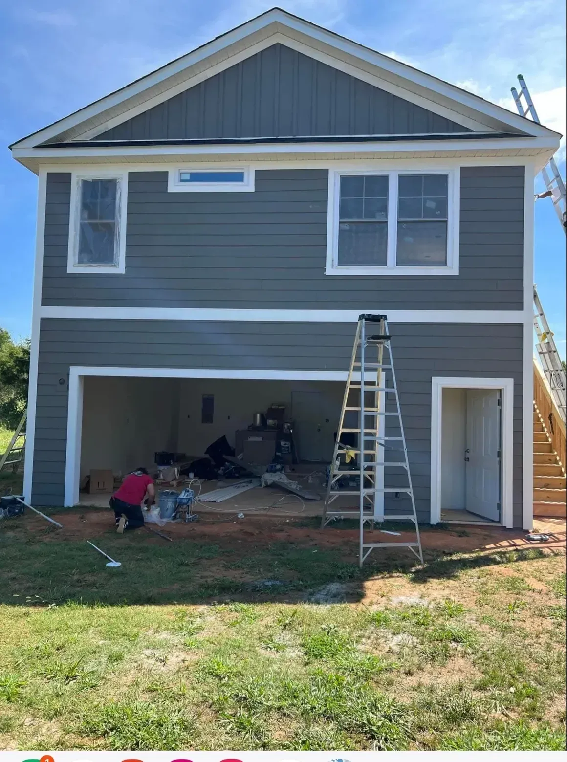A person works inside a gray-sided house with a large open garage door and a ladder set up on the lawn.