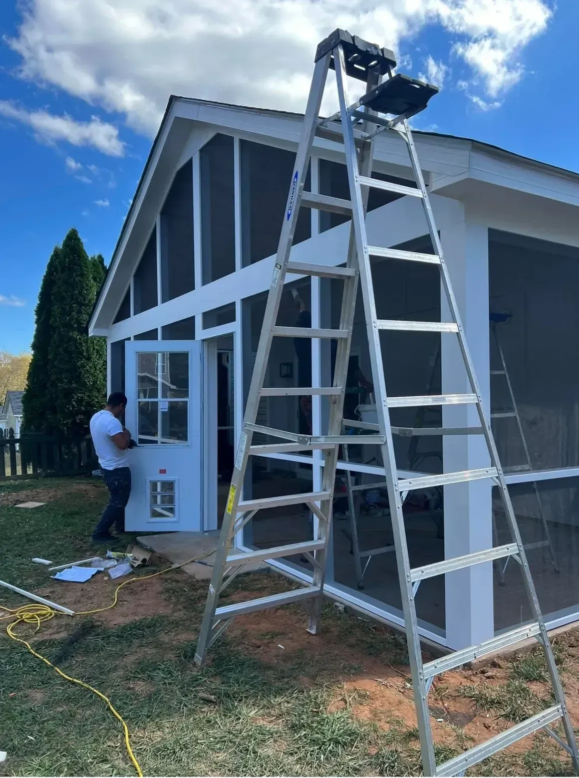 A person installs a screen door on a white porch structure with a tall metal ladder in the foreground.