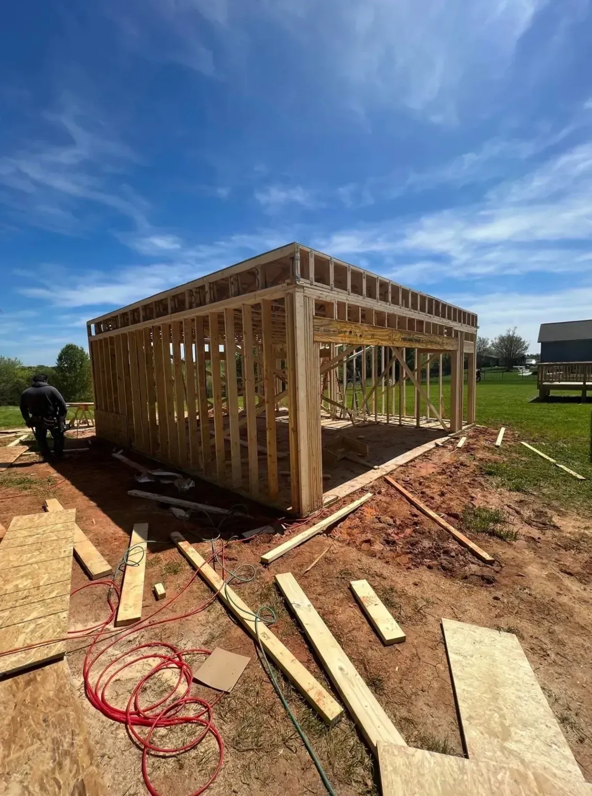 The wooden frame of a building under construction sits on a dirt lot under a bright blue sky.