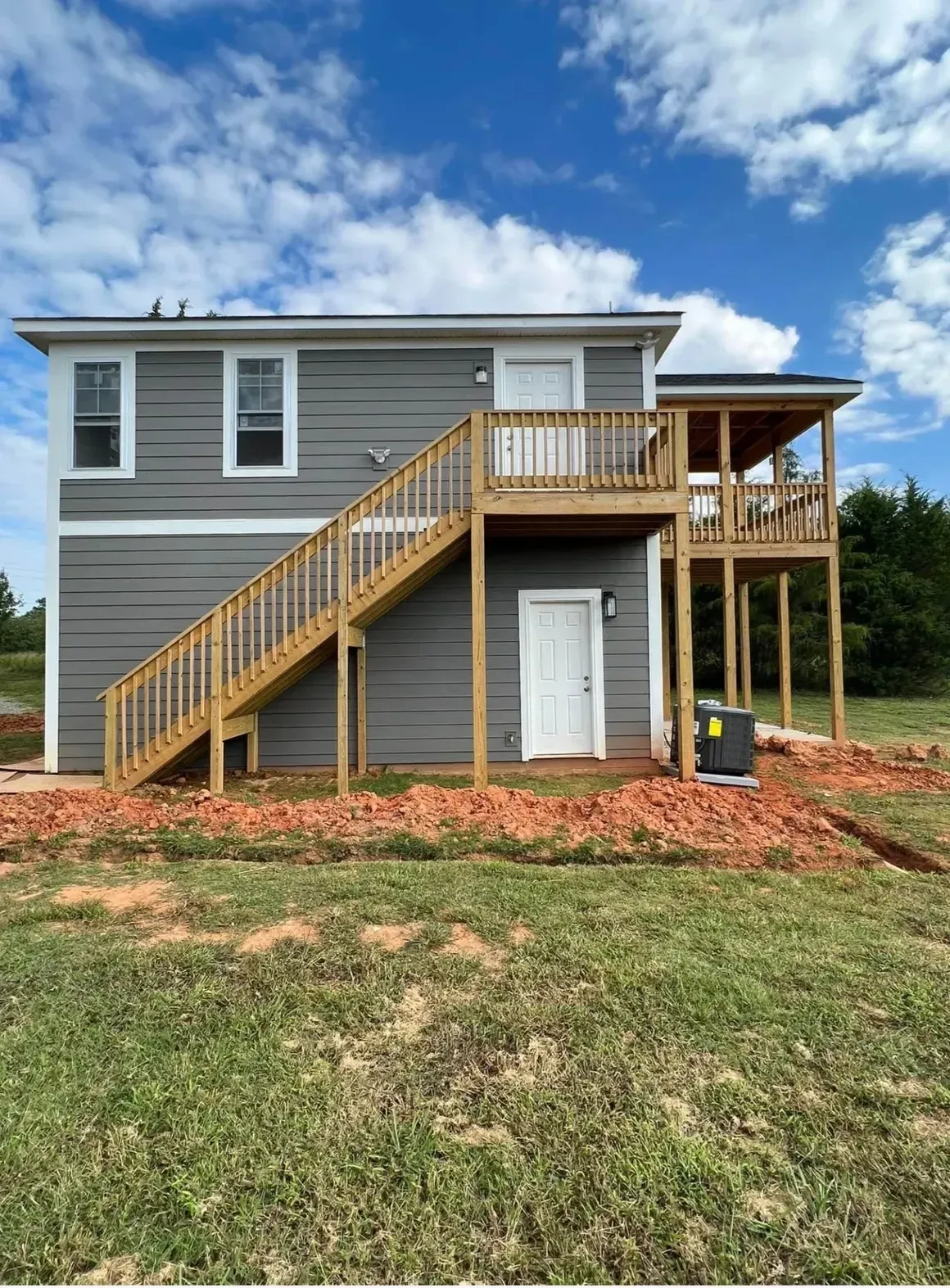 A two-story grey house with a wooden staircase leading to an upper deck, situated on a grassy lot with red soil.