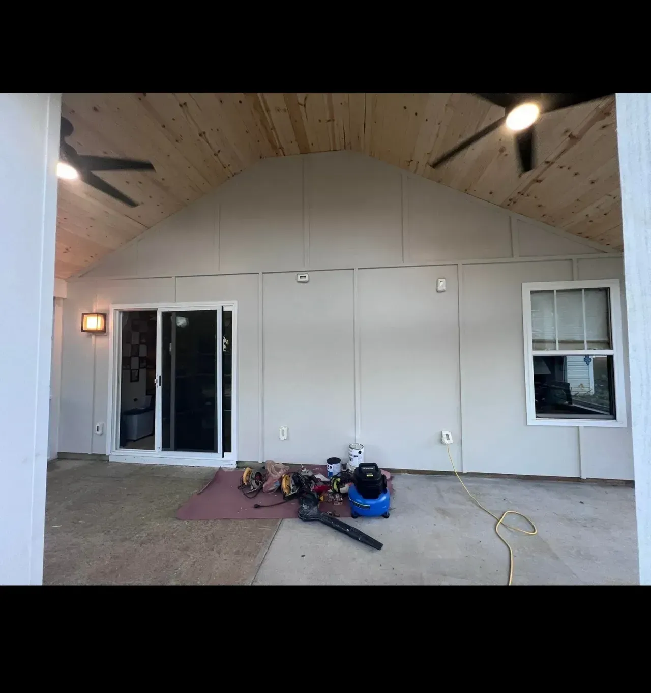 A covered outdoor patio featuring a wood-paneled vaulted ceiling, light-colored walls, a sliding glass door, and a window.