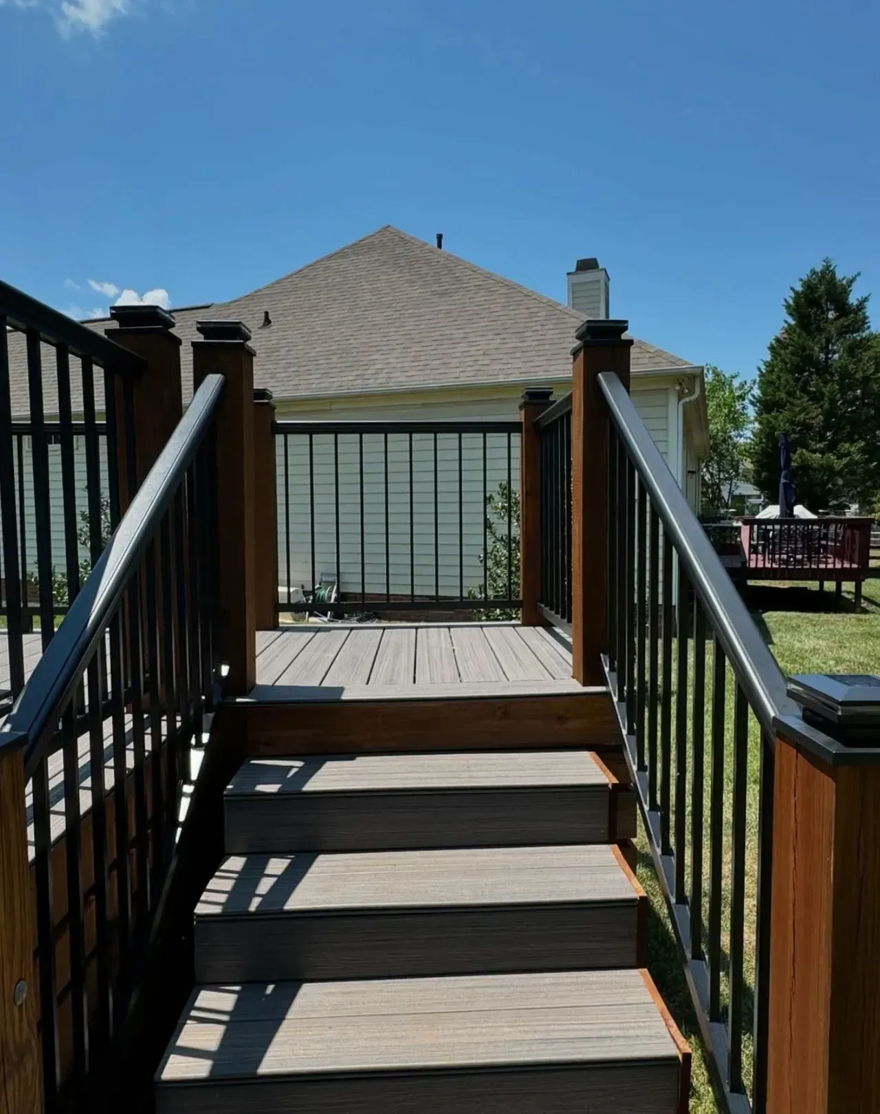 A set of wooden deck stairs with black railings leads up to a landing and a view of a suburban house under a blue sky.