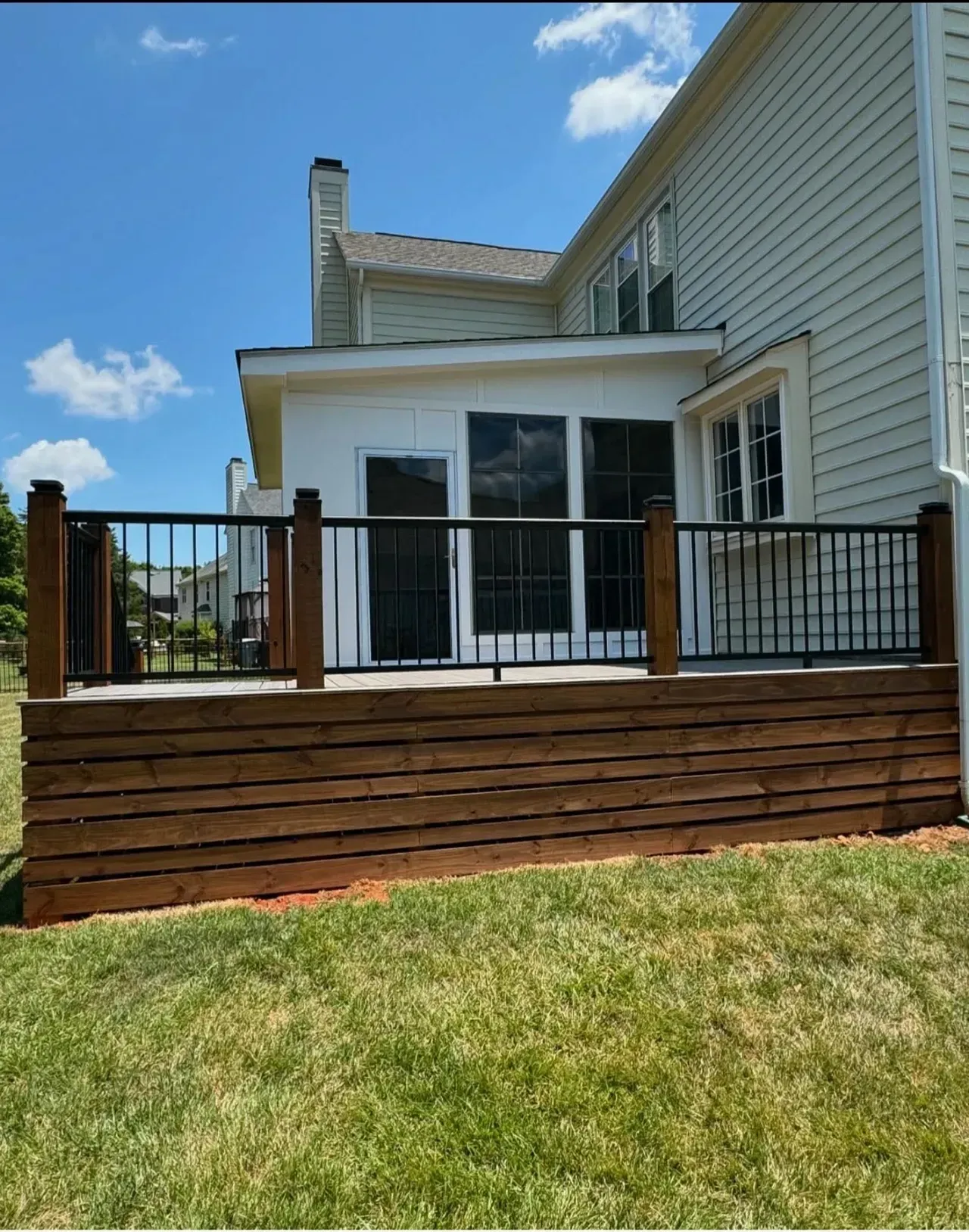 A backyard deck with wooden horizontal skirting and black metal railings attached to a light-colored two-story house.