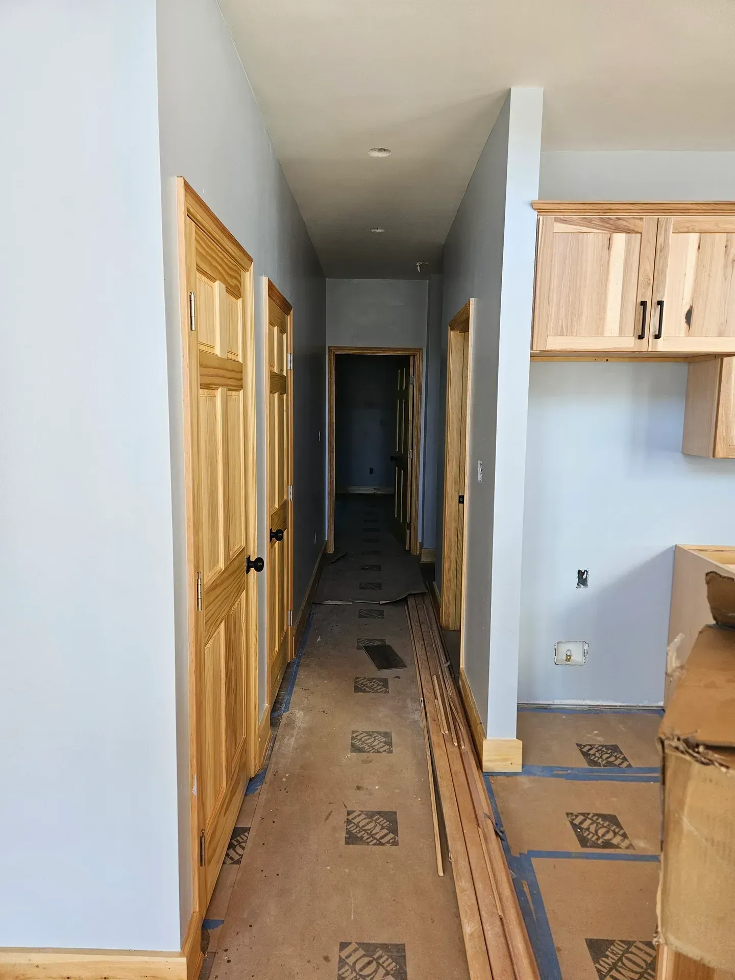 A hallway under construction with light blue walls, natural wood doors, and protective paper covering the floor.