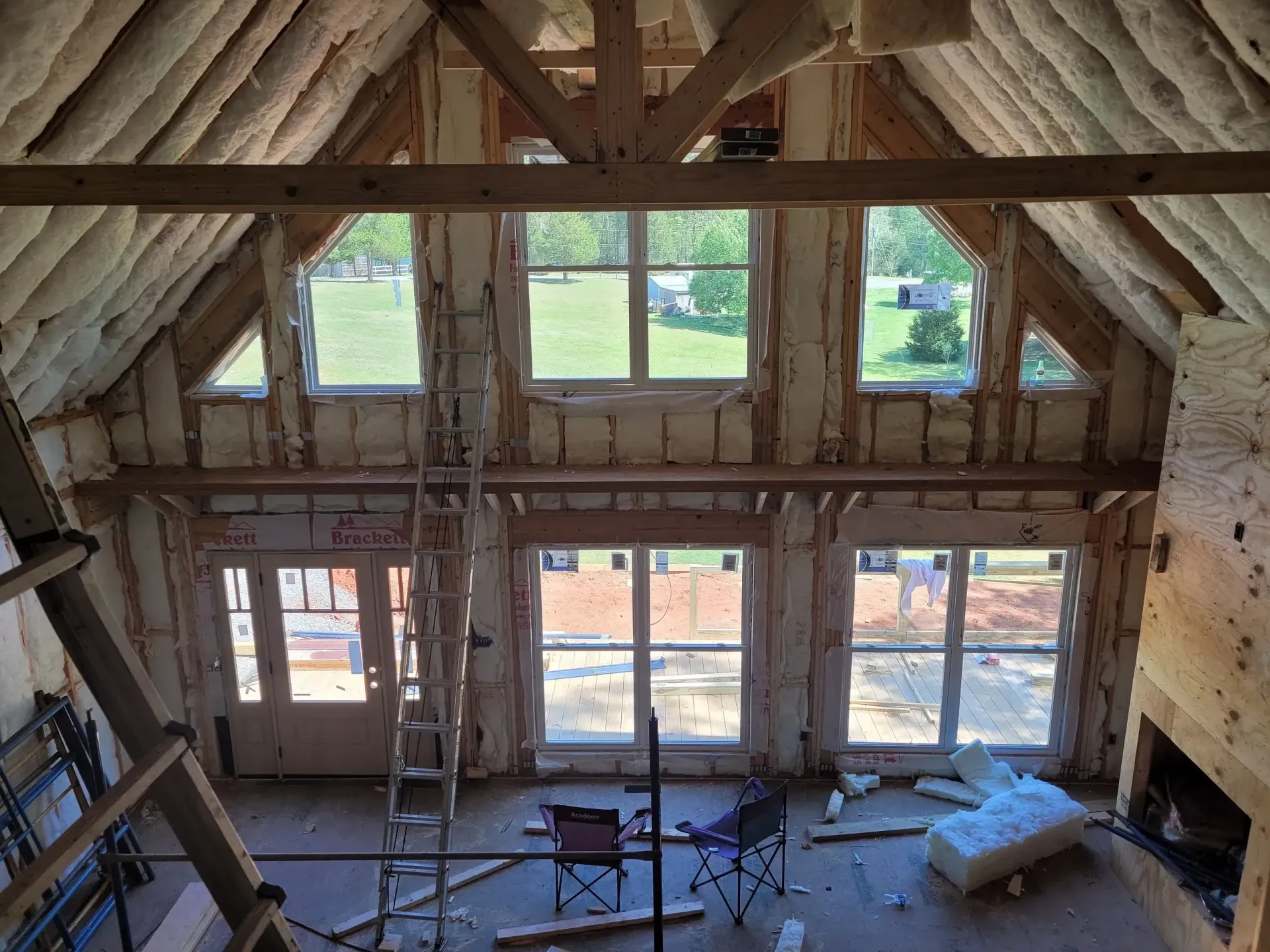 An unfinished room with a vaulted ceiling, exposed wooden beams, spray foam insulation, and large windows.