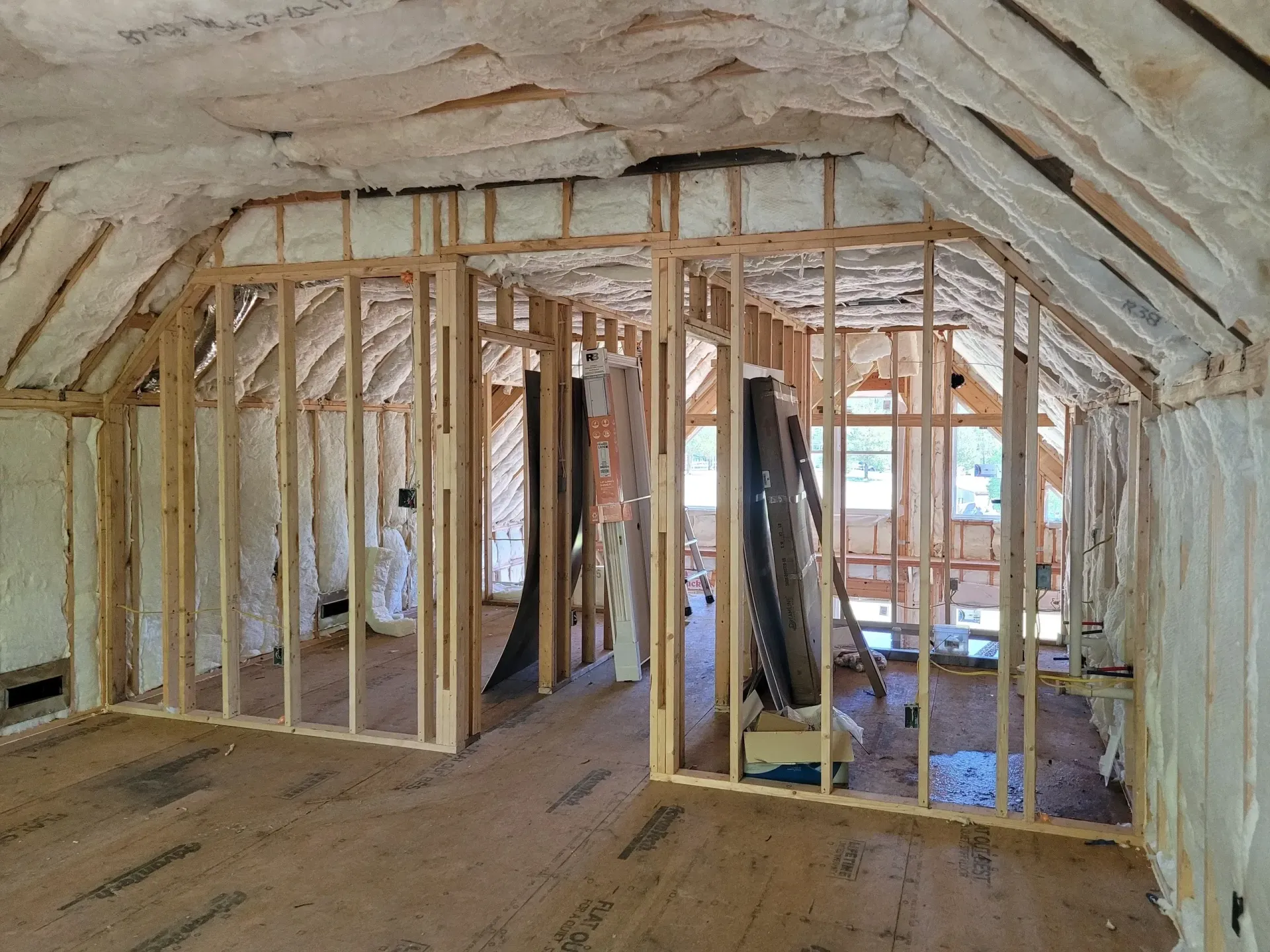 An attic space under renovation with exposed wood wall framing, unfinished flooring, and spray foam insulation.