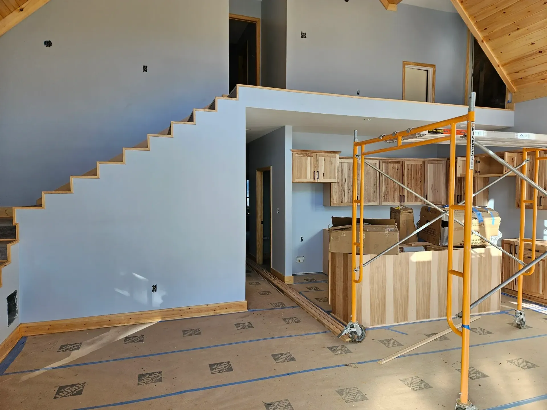A room under construction with a wooden staircase, light blue walls, wood cabinets, and yellow scaffolding.
