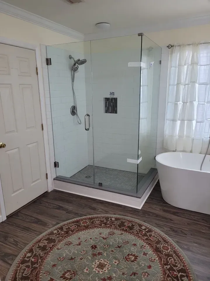 A modern bathroom with a glass-enclosed shower, a white freestanding tub, and a round patterned rug on a wood floor.