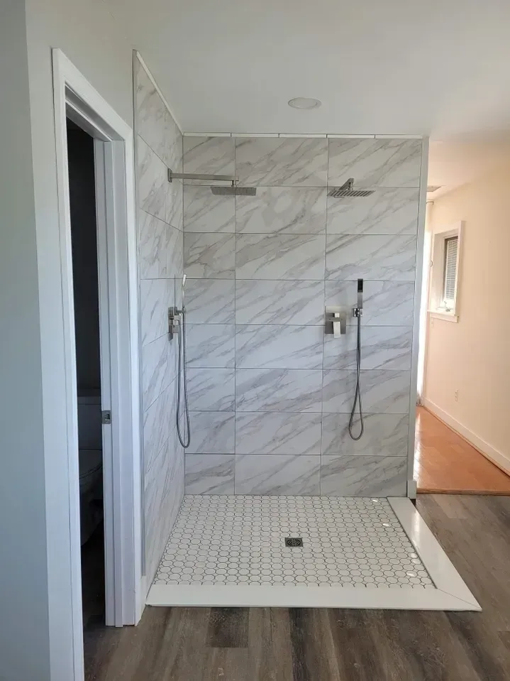 A walk-in shower with marble-look wall tiles, a hexagonal floor, and two chrome shower heads and handheld sprayers.
