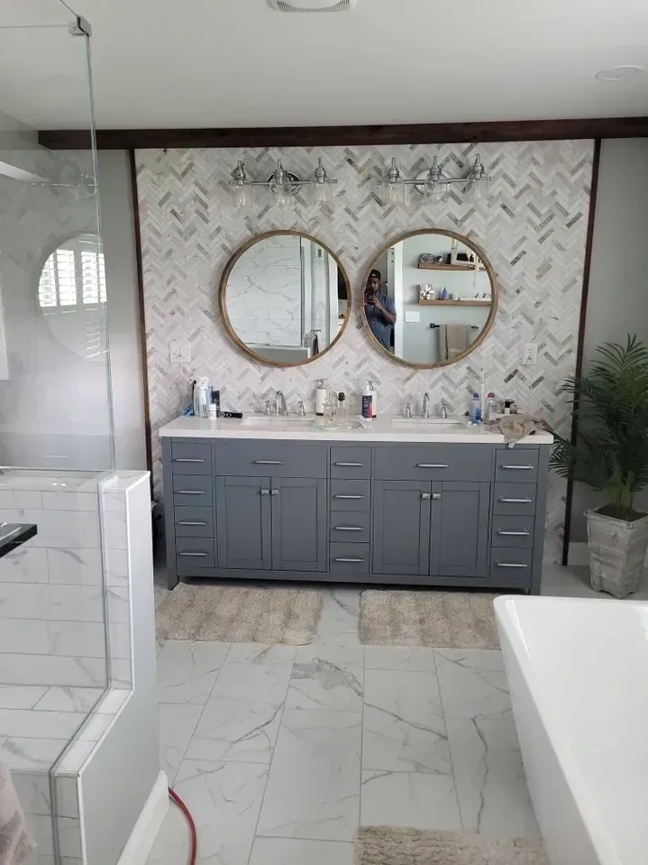 A modern bathroom with a double vanity, round mirrors, herringbone-patterned wall tile, and a glass-enclosed shower.