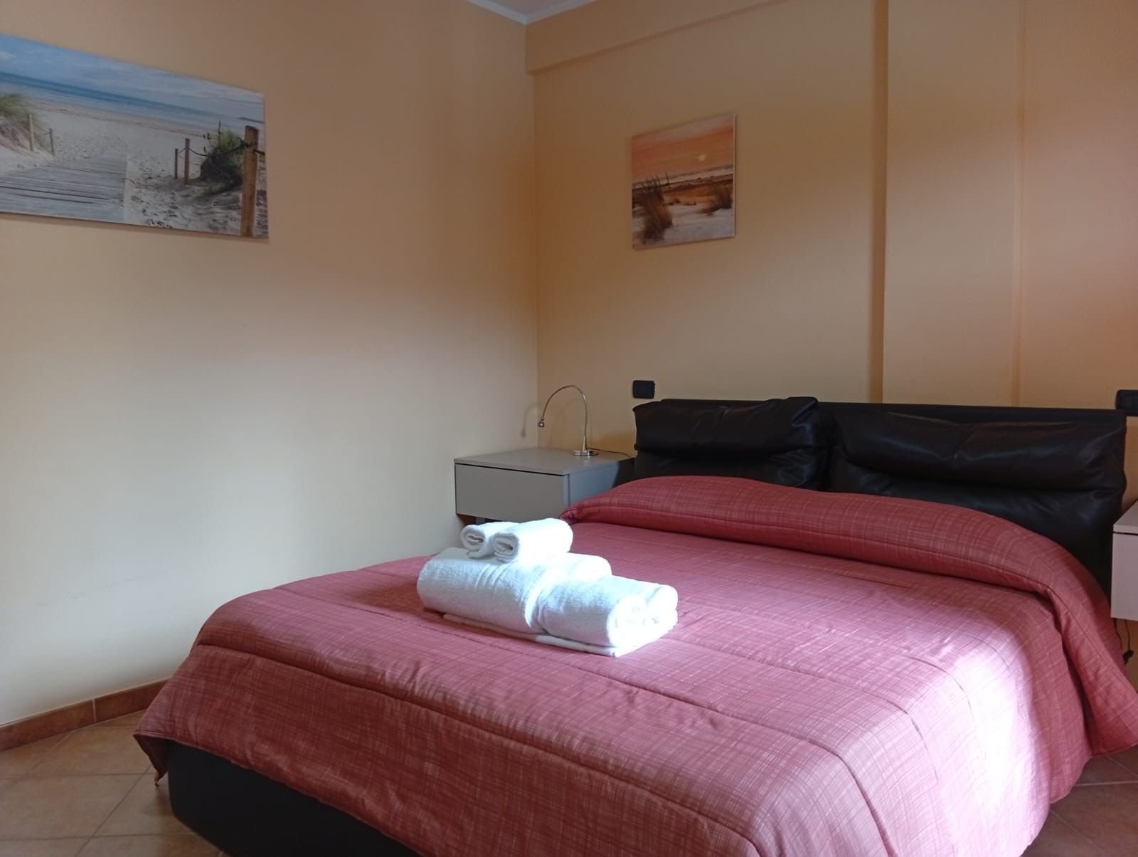 Camera da letto con letto matrimoniale, lampada da comodino e tavolino decorativo. Pareti bianche, pavimento marrone.