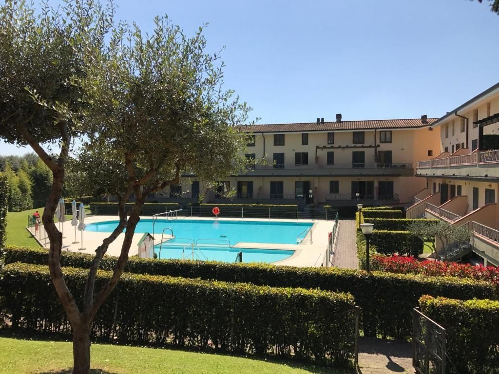 Piscina di fronte a un edificio a più piani. Alberi verdi e siepi circondano l'area della piscina nelle giornate di sole.
