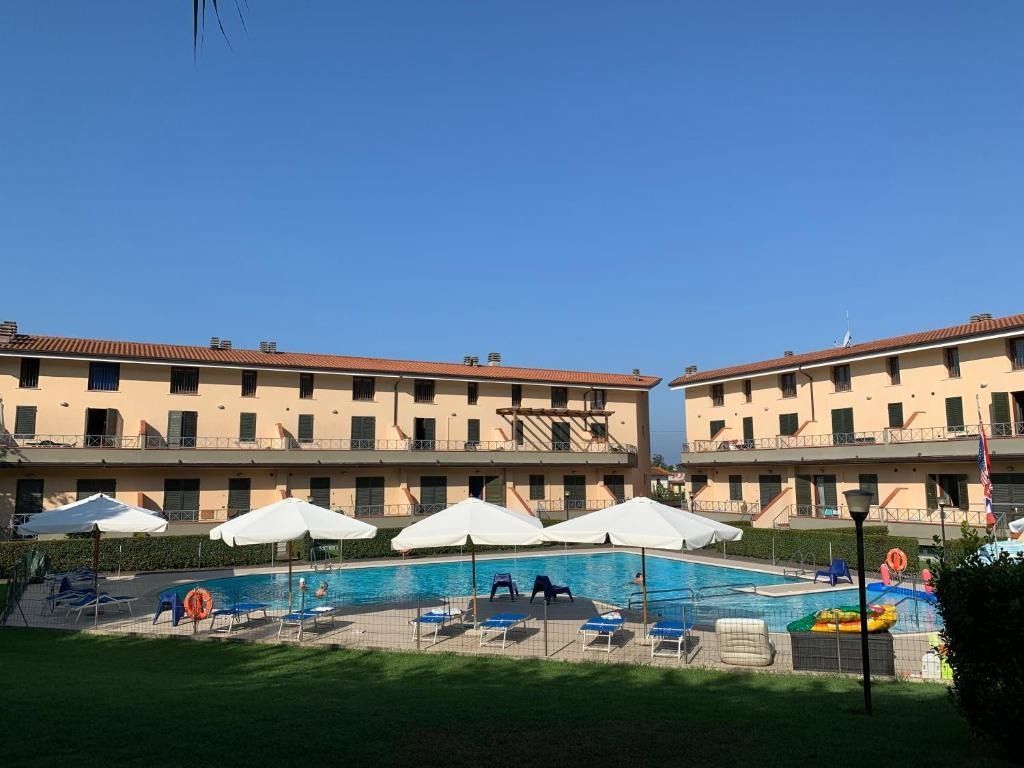Edifici a due piani fiancheggiano una piscina con ombrelloni e sedie a sdraio sotto un cielo limpido.