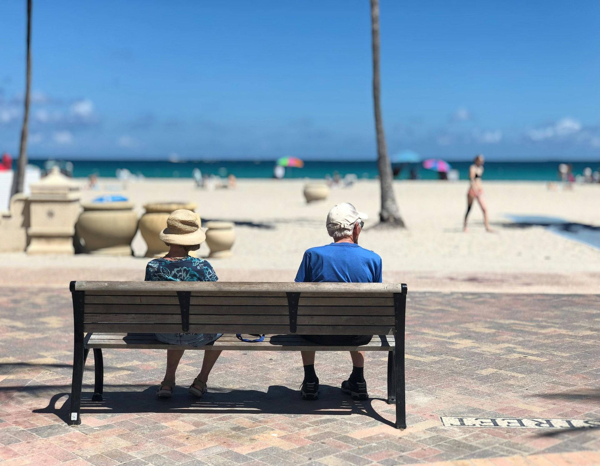 Two retired people sit on a bench, facing the beach on a sunny day.
