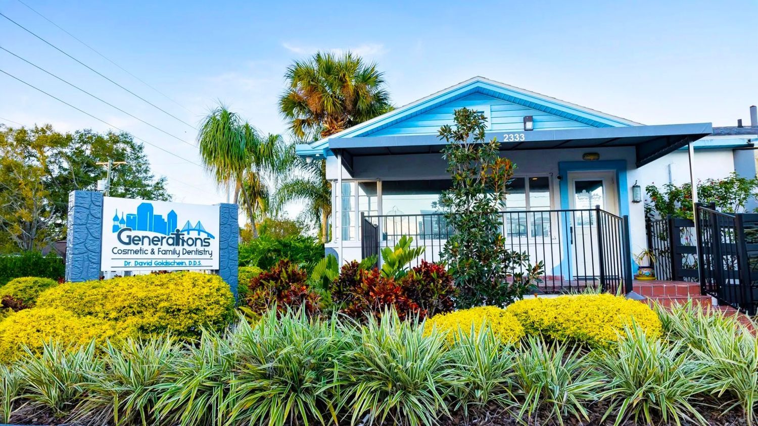A blue house with a sign in front of it surrounded by plants and bushes.